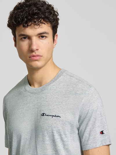 Męski model nosi szary t-shirt z mieszanki bawełny model 'ICONS CONTRAST' od CHAMPION z wyhaftowanym logo, nowoczesny krój, okrągły dekolt, długość tyłu w rozmiarze M: 70 cm, zbliżenie na górną część ciała.