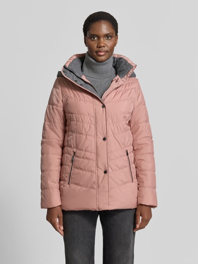 Modelka nosi 'Jacke in Stepp-Optik mit Kapuze' od FUCHS SCHMITT, kolor różowy, z efektem pikowania, taliowany krój, kaptur, ocieplana stójka, wpuszczane kieszenie z zamkiem błyskawicznym, długość tyłu w rozmiarze 36: 74 cm, szerokość ramion w rozmiarze 36: 40 cm, szerokość klatki piersiowej w rozmiarze 36: 48 cm, długość rękawów w rozmiarze 36: 64 cm, widok całej sylwetki z przodu