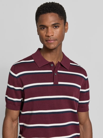 Männermodel trägt Regular Fit Poloshirt mit polokraag van Christian Berg Men van puur katoen in bordeaux met witte en blauwe strepen, knoopsluiting, ruglengte bij maat M 68 cm, mouwlengte 25 cm, nahaufnahme bovenlichaam.