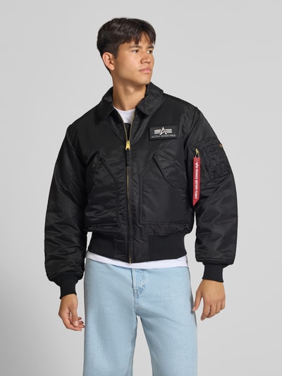 Männermodel trägt Bomberjacke mit seitlichen Pattentaschen und Label-Patch von Alpha Industries in Schwarz, Kentkragen, Reißverschluss, Regular Fit, Detailaufnahme Oberkörper.