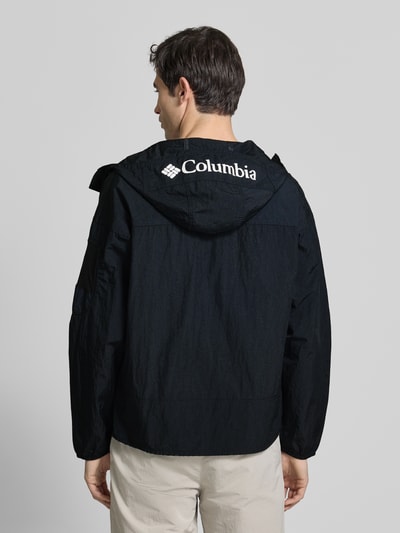 Männermodel trägt Windbreaker mit verdecktem Reißverschluss von Columbia, Schwarz, Regular Fit, Kapuze mit Tunnelzug, Logo, Ganzkörperansicht Rückansicht. Rückenlänge 70 cm, Schulterbreite 64 cm, Brustweite 57 cm, Ärmellänge 56 cm.