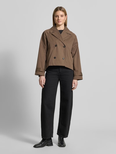 Frauenmodel trägt Regular Fit Trenchcoat-Jacke aus Baumwoll-Mix Modell 'NEW CILO' von Selected Femme in Optik: Unifarben mit Reverskragen, doppelreihiger Knopfleiste, Rückennaht mit Kellerfalte, Langarm, Raglanärmel, Riegel am Ärmelabschluss, Innentasche und seitlichen Leistentaschen, Textur glatt, Farben braun und schwarz, Ganzkörperansicht von vorne.