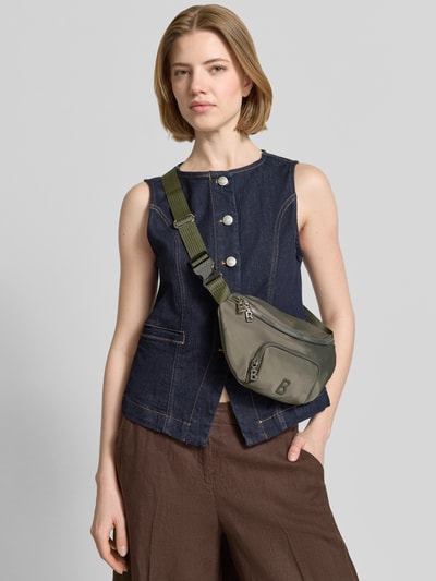 Frauenmodel trägt olivgrüne Handtasche mit Label-Detail Modell 'verbier play 1.0 janica' von BOGNER, Zweiwege-Reißverschluss, Nahaufnahme Oberkörper.