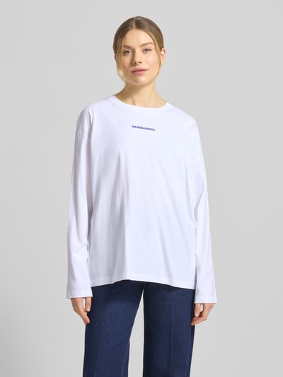Kobiece modelka nosi Oversized Langarmshirt aus reiner Baumwolle Modell 'FIETAA SMILE' od Armedangels, biała gładka dzianina, logo na piersi, długi rękaw, widok całej sylwetki z przodu.