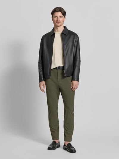 Męski model nosi Slim Fit Hose mit Paspeltaschen am Gesäß od CRUNA, materiał z domieszką stretchu, krój slim fit, kieszenie wpuszczane, oliwkowy kolor, szerokość w rozmiarze 50: 44 cm, długość nogawki wewnętrznej w rozmiarze 50: 73 cm, widok całej sylwetki z przodu.