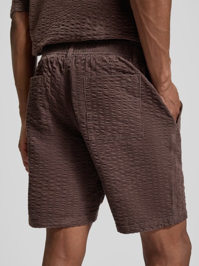 Mężczyzna nosi Szorty Straight Fit mit Strukturmuster od JAKE*S STUDIO MEN, mieszanka bawełny, fakturowany wzór, wpuszczane kieszenie, zapięcie na guzik i zamek błyskawiczny, szerokość w rozmiarze M: 45 cm, długość wewnętrzna nogawki w rozmiarze M: 20 cm, brązowe, widok całej sylwetki z tyłu.