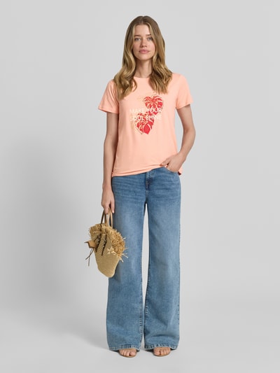 Frauenmodel trägt Damen T-Shirt mit Statement- und Motiv-Print Modell 'RINA' von Soyaconcept, Reine Baumwolle, Regular-Fit, Rundhalsausschnitt, lichte Pfirsichfarbe und rote Akzente, kombiniert mit blauer Jeans, Rückenlänge bei Größe S: 60 cm, Ganzkörperansicht von vorne.