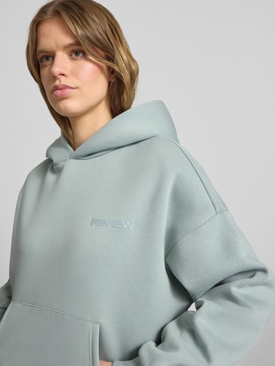 Frauenmodel trägt Hoodie mit Logo und Känguru-Tasche von Review, Damen, Baumwoll-Mix, Relaxed-Fit, Logo, Känguru-Tasche, Nahaufnahme Oberkörper.