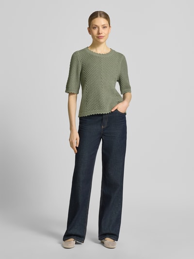 Frauenmodel trägt Strickshirt mit 1/2-Arm Modell 'Carla' von mbyM aus Material-Mix mit Baumwoll-Anteil im Grobstrick, Gerader-Schnitt, Rundhalsausschnitt, dezentes Grün, grobe Textur, Ganzkörperansicht von vorne.