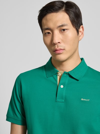Męski model nosi Regular Fit Poloshirt mit Logo-Stitching od Gant, zielony kolor, elastyczna bawełna, krój regular fit, kołnierzyk polo, krótkie rękawy, zbliżenie na górną część ciała.