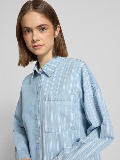 Modelka kobieca nosi Oversized Jeansbluse mit Brusttasche Modell 'KATRI' od Noisy May, jasnoniebieski kolor, bawełna, krój oversized, kieszeń na piersi, kołnierzyk koszulowy, listwa guzikowa, ramiona opuszczone, mankiety sportowe, zaokrąglony dół, długość pleców w rozmiarze S 72 cm, długość rękawa w rozmiarze S 50 cm, tekstura miękkiego denimu, widok z przodu, zbliżenie na górną część ciała.