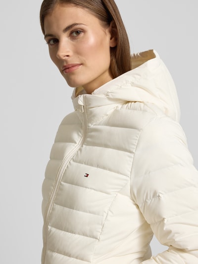 Kobiece model prezentuje Slim Fit Daunenjacke mit abnehmbaren Kapuze od Tommy Hilfiger, efekt: jednolity kolor, krój slim fit, odpinany kaptur i stójka, zamek błyskawiczny, wyhaftowane logo, długie rękawy z elastycznym wykończeniem, zaokrąglone zakończenie, hydrofobowa, boczne kieszenie na zamek, detal z logo marki, jasna kolorystyka, tekstura pikowana. Zbliżenie na górną część sylwetki – detalowa perspektywa.