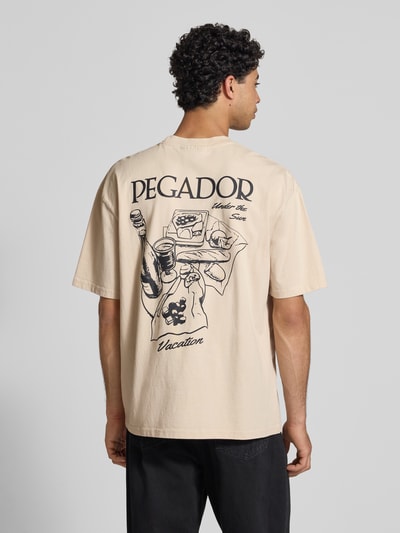 Männermodel trägt Oversized T-Shirt mit Label-Print Modell 'Paide' von Pegador in beige, pure katoen, rugprint, Ganzkörperansicht Rückansicht.