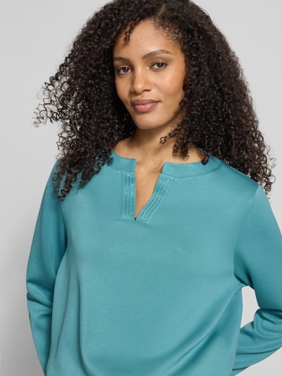 Frauenmodel trägt Regular Fit Sweatshirt mit Viskose-Anteil von Tom Tailor, Optik: Unifarben, Applikation: Label-Stitching-Applikation, Strasssteine, V-Ausschnitt, Ärmelform: Langarm, Ärmelabschluss und Rumpfabschluss: elastisch, Materialverarbeitung: Sweat, Farbe türkis, glatte Textur, Nahaufnahme Oberkörper.