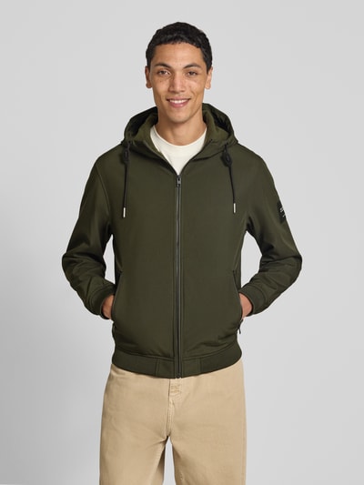 Männermodel trägt Regular Fit Jacke mit Logo-Detail von JACK & JONES in Olivgrün, Kapuze, Softshell, seitliche Reißverschlusstaschen, Ganzkörperansicht von vorne.