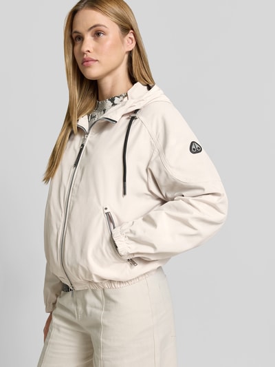 Frauenmodel trägt Jacke mit Zweiwege-Reißverschluss von Moose Knuckles in Beige mit Kapuze, Tunnelzug, Relaxed-Fit, Eingrifftaschen mit Reißverschluss, Rückenlänge 56 cm, Brustweite 56 cm, Ärmellänge 44 cm, Ganzkörperansicht von vorne.