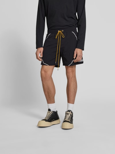 Männermodel trägt Shorts mit Label-Stitching von RHUDE, schwarze Farbe, gelber Tunnelzug, weiße Details, Eingrifftaschen, Ganzkörperansicht von vorne.