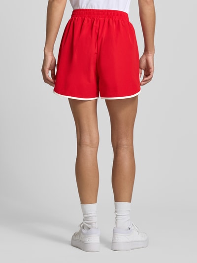Frauenmodel trägt Track Shorts mit elastischem Bund und Label-Detail von Karo Kauer in Rot, Material-Mix mit Stretch-Anteil, Loose-Fit, Ganzkörperansicht Rückansicht. Bundweite bei Größe S: 36 cm, Innenbeinlänge bei Größe S: 9 cm.