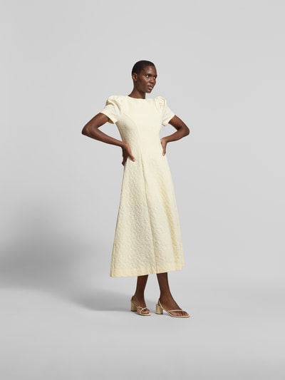 Frauenmodel trägt Midikleid mit floralem Allover-Detail von ROTATE in Gelb, Elasthan-Mix, taillierte Passform, A-Linie, kurzärmelig, Ganzkörperansicht von vorne.