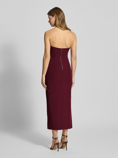 Vrouwenmodel draagt midi-jurk in bordeauxrood materiaalmix met stretch, recht model, loopsplit, cut-out detail, model 'ELENI' van Bardot, ruglengte maat 36: 108 cm, borstwijdte: 40 cm, ritssluiting achter, off-shoulder-look, volledige lichaamsaanzicht achteraan.