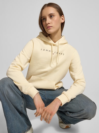 Modelka kobieca ma na sobie Regular Fit Hoodie aus Baumwoll-Mix od Tommy Jeans, jednolity kolor, nadruk z logo, krój regular fit, kapuzę z troczkiem, długie rękawy i ściągacze, materiał bawełniany typu sweat, widok na cały korpus z przodu.