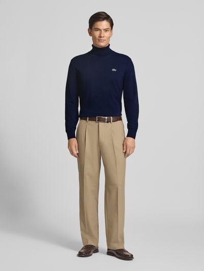 Männermodel trägt Regular Fit Pullover aus reiner Baumwolle von Lacoste, dunkelblau, kombiniert mit beiger Stoffhose, Ganzkörperansicht von vorne, Herren Turtleneck-Pullover von Lacoste.