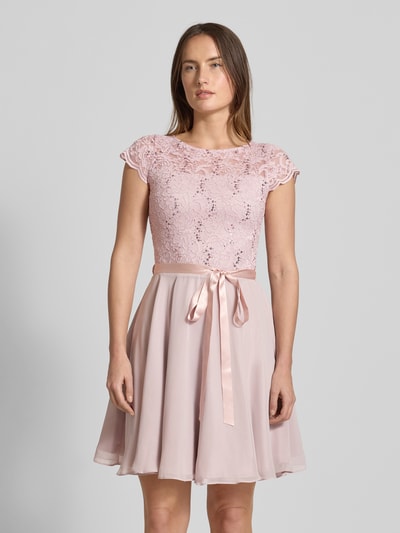 Modelka kobieca nosi Cocktailkleid mit Spitzenbesatz und Bindegürtel od SWING; różowy materiał, koronka, cekiny, krótka A-linia, ujęcie całego ciała z przodu.