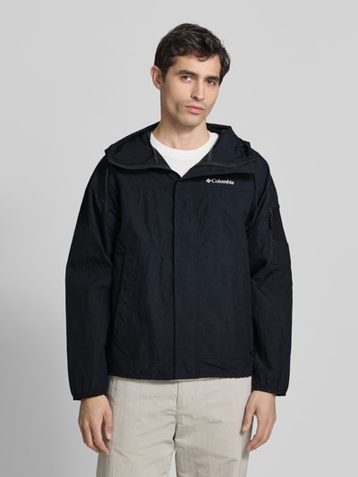 Männermodel trägt Windbreaker mit verdecktem Reißverschluss von Columbia in Schwarz mit Logo, Regular-Fit, Ganzkörperansicht von vorne, Rückenlänge 70 cm, Schulterbreite 64 cm, Brustweite 57 cm, Ärmellänge 56 cm, Kapuze mit Tunnelzug, glatte Textur, schlichte Gestaltung.