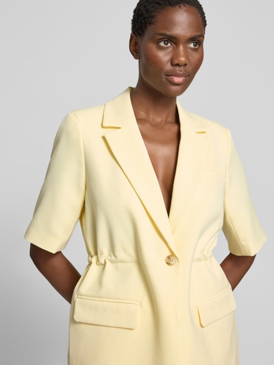 Kobieta modelka ma na sobie żakiet Relaxed Fit Blazer mit Viskose-Anteil Model 'Likka' od YAS, elastyczna mieszanka materiałów z dodatkiem wiskozy, kołnierz z połami, rękaw o dł. 1/2, zapięcie na guzik, kieszenie z patką, długość tyłu w rozmiarze 36: 70 cm, jasny żółty kolor, gładka tekstura, detaliczna faktura materiału, nahaufnahme górnej części ciała.