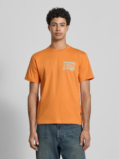 Męski model nosi Regular Fit T-Shirt mit Statement-Print od Replay z czystej bawełny, krój regular fit, okrągły dekolt, pomarańczowy kolor, długość tyłu w rozmiarze M: 62 cm, całkowity widok z przodu.