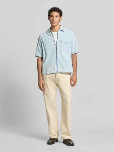 Männermodel trägt Regular Fit Jeanshemd aus reiner Baumwolle Modell 'CAMICIA' von Armani Exchange in Hellblau, Ganzkörperansicht von vorne, Kurzarm, Kentkragen, Zweiwege-Reißverschluss, sichtbarer Brusttasche und dezentem Brand-Stitching, kombiniert mit heller Hose, glatte Textur.