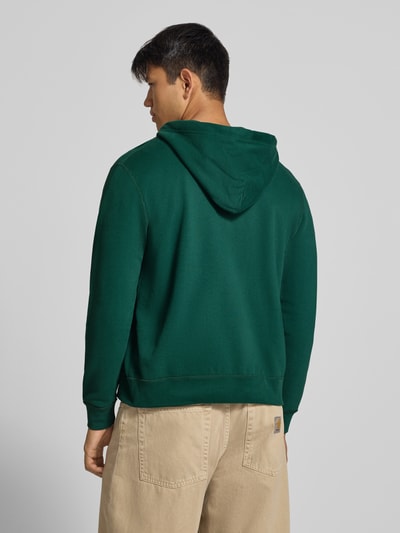 Männermodel trägt Regular Fit Sweatjacke aus Baumwoll-Mix mit Label-Detail von Superdry in Dunkelgrün, Rückenansicht, Ganzkörperansicht Rückansicht. Produktmerkmale: Kapuze mit Tunnelzug, getrennte Känguru-Tasche, elastische Abschlüsse, Rückenlänge 65 cm, Ärmellänge 65 cm.
