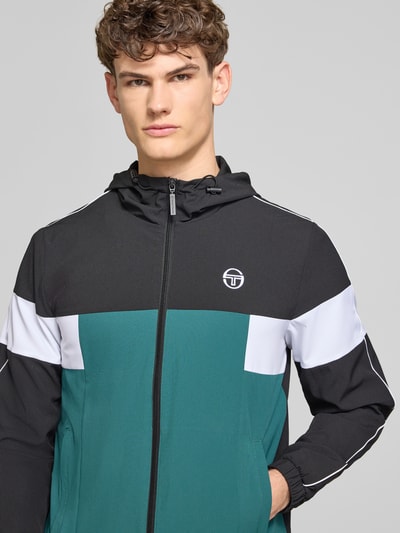 Männermodel trägt Regular Fit Jacke mit Logo-Stitching Modell 'Caspio' von SERGIO TACCHINI, schwarz-grün-weiß, Nahaufnahme Oberkörper. Elastischer Material-Mix, Kapuze mit Tunnelzug, Reißverschluss, Eingrifftaschen und elastische Ärmelabschlüsse sichtbar.