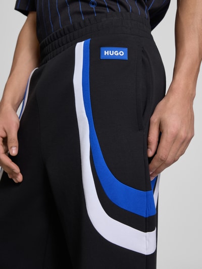 Männermodel trägt Relaxed Fit Sweatpants aus Baumwoll-Mix Modell 'Nalotron' von Hugo Blue mit blauen und weißen Kontraststreifen, Nahaufnahme Hüfte bis Oberschenkel.