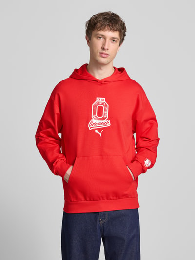 Männermodel trägt Relaxed Fit Hoodie mit Känguru-Tasche von PUMA PERFORMANCE in Rot mit weißem Label-Detail, Ganzkörperansicht von vorne.