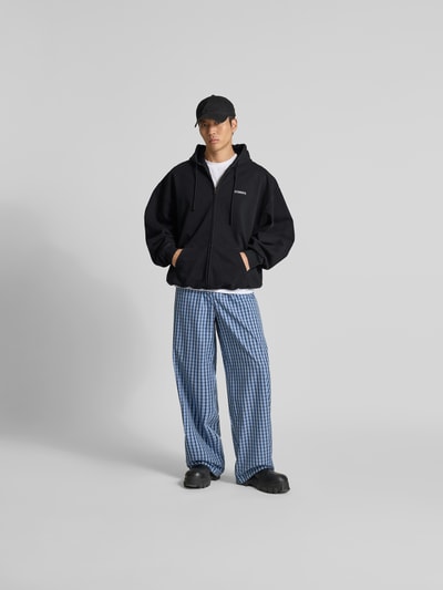 Männermodel trägt Oversized Sweatjacke aus Baumwolle von VETEMENTS, schwarz, mit Zweiwege-Reißverschluss, Känguru-Tasche, elastische Abschlüsse, kombiniert mit blau karierten Hosen, Ganzkörperansicht von vorne. Rückenlänge 75 cm, Schulterbreite 50 cm, Ärmellänge 78 cm bei Größe M.