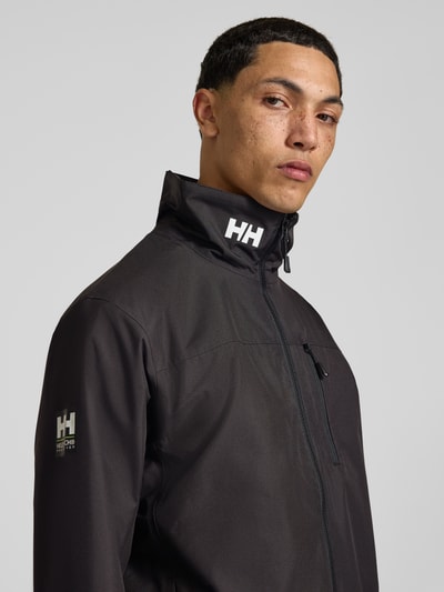 Männermodel trägt Regular Fit Jacke mit Stehkragen Modell 'Crew' von Helly Hansen in Schwarz, Logo-Print, Nahaufnahme Oberkörper.