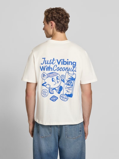 Männermodel trägt Regular Fit T-Shirt aus reiner Baumwolle Modell 'VIBING COCO' von REVIEW mit blauem Print auf Rückseite, Ganzkörperansicht Rückansicht, Herren T-Shirt von REVIEW x ANSON´S, Rückenlänge bei Größe M 68 cm, Print zeigt Illustrationen und Schrift.