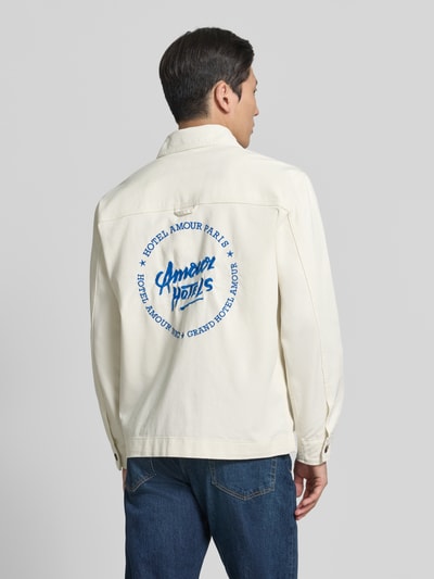 Męski model nosi Regular Fit Jeansjacke aus Bio-Baumwoll-Mix Modell 'HOTEL AMOUR' od SELECTED HOMME, kremowy kolor, nadruk na plecach, krój regular fit, długie rękawy, klasyczny kołnierz, widok całej sylwetki z tyłu.