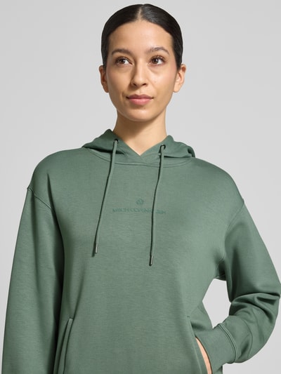 Modelka kobieca ma na sobie Hoodie z tunelem model 'IMA' od MSCH Copenhagen, kolor zielony, miks wiskozy, prosty krój, z kapturem, kieszenie boczne, zbliżenie na górną część ciała z przodu.
