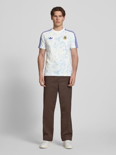 Männermodel trägt Regular Fit Trikot aus reiner Baumwolle von Adidas Originals mit Optik:Allover-Muster, Regular-Fit, Logo-Stitching, Kontraststreifen, Rundhals, Kurzarm, Materialverarbeitung: Jersey, Ganzkörperansicht von vorne.
