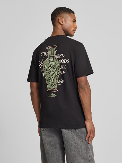 Mężczyzna nosi T-Shirt mit rückseitigem Motiv-Print Modell 'Ben Faience' von Les Deux, czarny bawełniany, prosty krój, nadruk statement oraz label na plecach, widok całej sylwetki z tyłu.