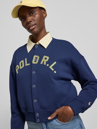 Vrouwenmodel draagt Sweatjacke mit Label-Print und Knopfleiste van Polo Ralph Lauren in katoenmix, relaxed fit, ronde hals, blauwe kleur, borstprint en knoopsluiting, detailopname bovenlichaam.