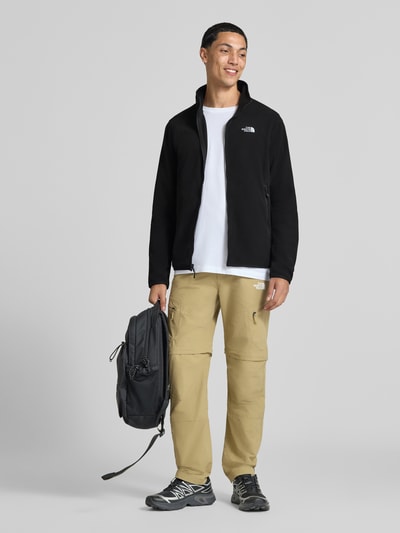 Męski model nosi kurtkę z polaru z zamkiem błyskawicznym Model 'GLACI' od The North Face, krój taliowany, kieszenie zapinane na zamek błyskawiczny, wyhaftowane logo, Długość tyłu w rozmiarze M: 66 cm, Długość rękawów w rozmiarze M: 68 cm, kolor czarny, widok całej sylwetki z przodu.