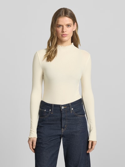 Modelka nosi Slim Fit Langarmshirt aus Lyocell-Mix von Marc O'Polo Denim, efekt: jednolity kolor, krój slim fit, wzór w paski, aplikacja: naszywka z logo, wyhaftowane logo, dekolt: wysokie zapięcie, rękawy: długi rękaw, zakończenie rękawów: falbanka, wykończenie: falbanka, wykończenie materiału: wykończenie materiału, materiał: mieszanka lyocellu, kolor jasnokremowy, struktura prążkowana. Ganzkörperansicht z przodu.