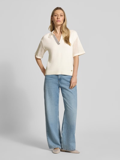 Modelka nosi Relaxed Fit Poloshirt in Strick-Optik mit Strasssteinbesatz od LIU JO SPORT, bawełna, krój relaxed fit, obszycie z kamieni stras, imitacja dzianiny, z krótkim rękawem, perforowanie, długość tyłu w rozmiarze S: 63 cm, długość rękawów w rozmiarze S: 22 cm, kolor ecru, luźna forma, dzianinowa tekstura, widok całej sylwetki z przodu.