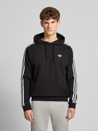 Männermodel trägt Hoodie mit Kapuze von adidas Originals in Schwarz, lockerer Schnitt, weiße Streifen, Känguru-Tasche, Ganzkörperansicht von vorne.
