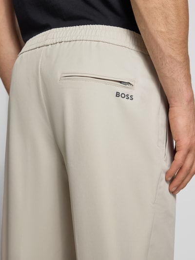 Mężczyzna nosi Tapered Fit Stoffhose mit Tunnelzug Modell 'T_FLEX' von BOSS Green, jasny beż, gładka faktura, elastyczny pas, średni stan, tylna kieszeń z zamkiem, label, boczne kieszenie, perspektywa: zbliżenie na biodra i tył.