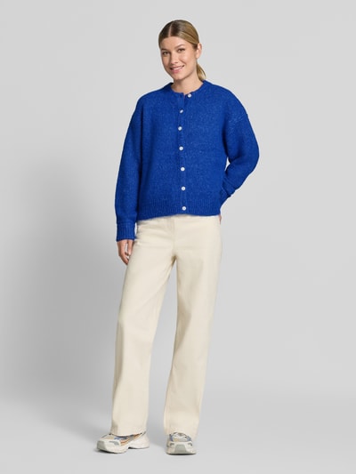 Kobiece modelka nosi kardigan rozpinany Relaxed Fit Strickjacke mit Woll-Anteil Modell 'Belablue' od ICHI, elastyczna mieszanka materiałów z dodatkiem wełny, pełna listwa guzikowa, krój z długim rękawem, okrągły dekolt, opuszczone ramiona, prążkowane ściągacze, długość tyłu w rozmiarze S: 57 cm, długość rękawów w rozmiarze S: 71 cm, jasne spodnie, biała i niebieska kolorystyka, gładka faktura, widok całej sylwetki z przodu.