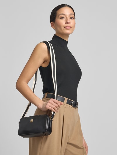 Frauenmodel trägt Umhängetasche in Leder-Optik von Tommy Hilfiger, einfarbig schwarz mit Logo und Schulterriemen, Nahaufnahme Oberkörper.